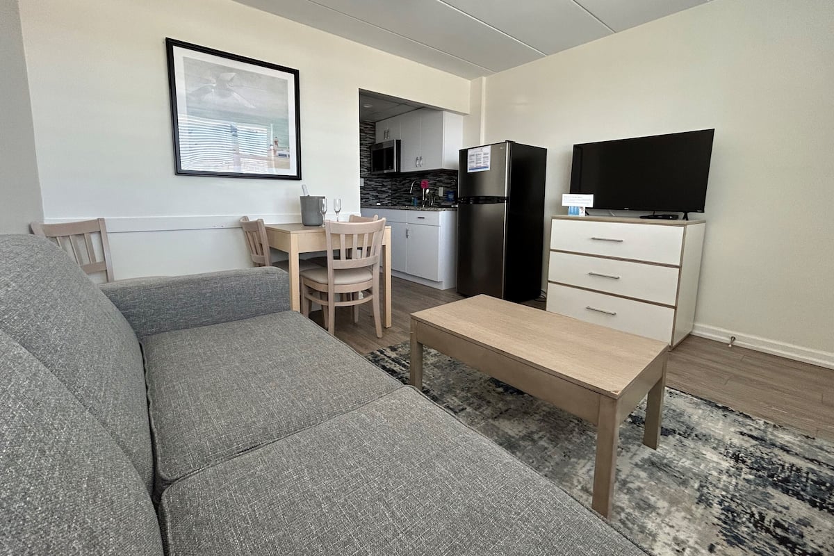 A modern living room with a grey sofa, a wooden dining table with chairs, a small TV on a cabinet, and a kitchen in the background.