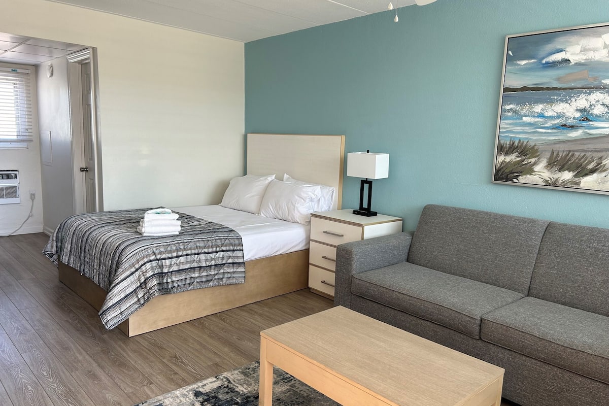 A bedroom with a bed, bedside table, lamp, and a painting of a beach scene.