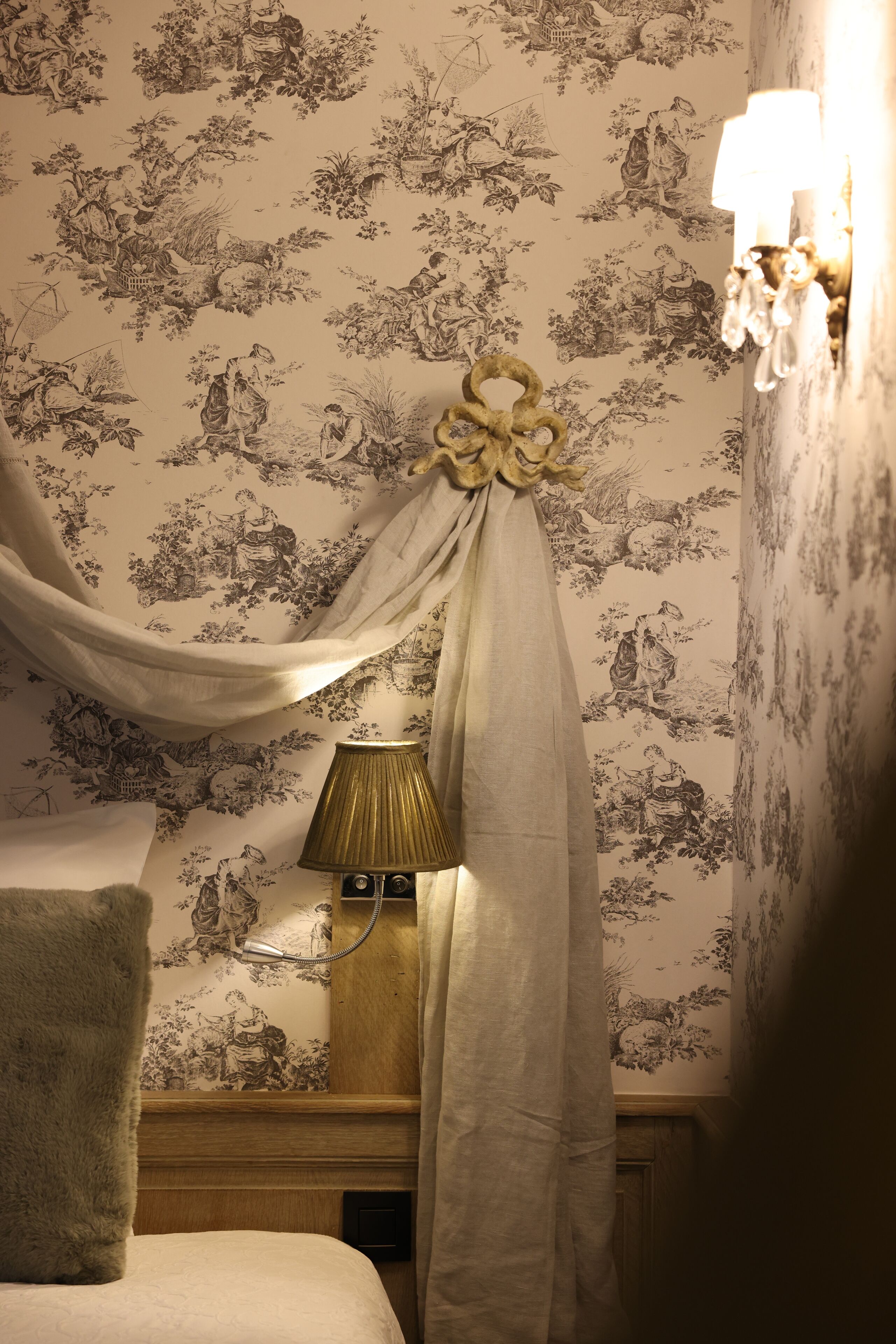 Een slaapkamer met bloemenbehang, een bed met een witte deken, een bedlampje en een nachtkastje van hout.