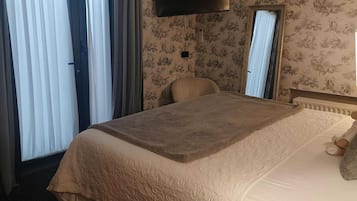 A hotel room with a large bed, a flat-screen TV mounted on the wall, a mirror, and a window with curtains.