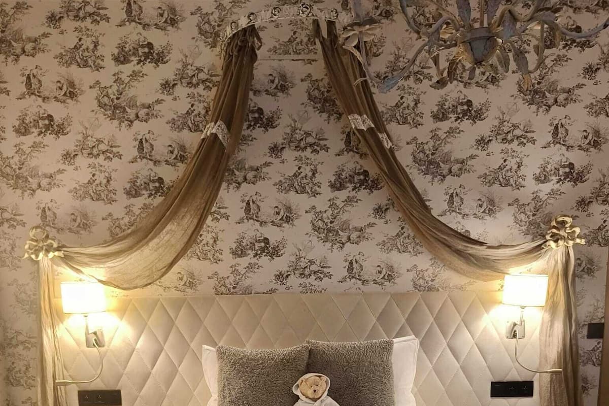 Een slaapkamer met een geborduurd hoofdbord, een zachte deken, een teddybeer op een kussen en een kroonluchter.