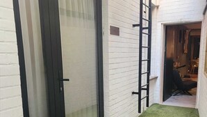 Un étroit couloir extérieur doté d’une porte coulissante en verre, d’un gazon synthétique vert et d’une échelle appuyée contre le mur.