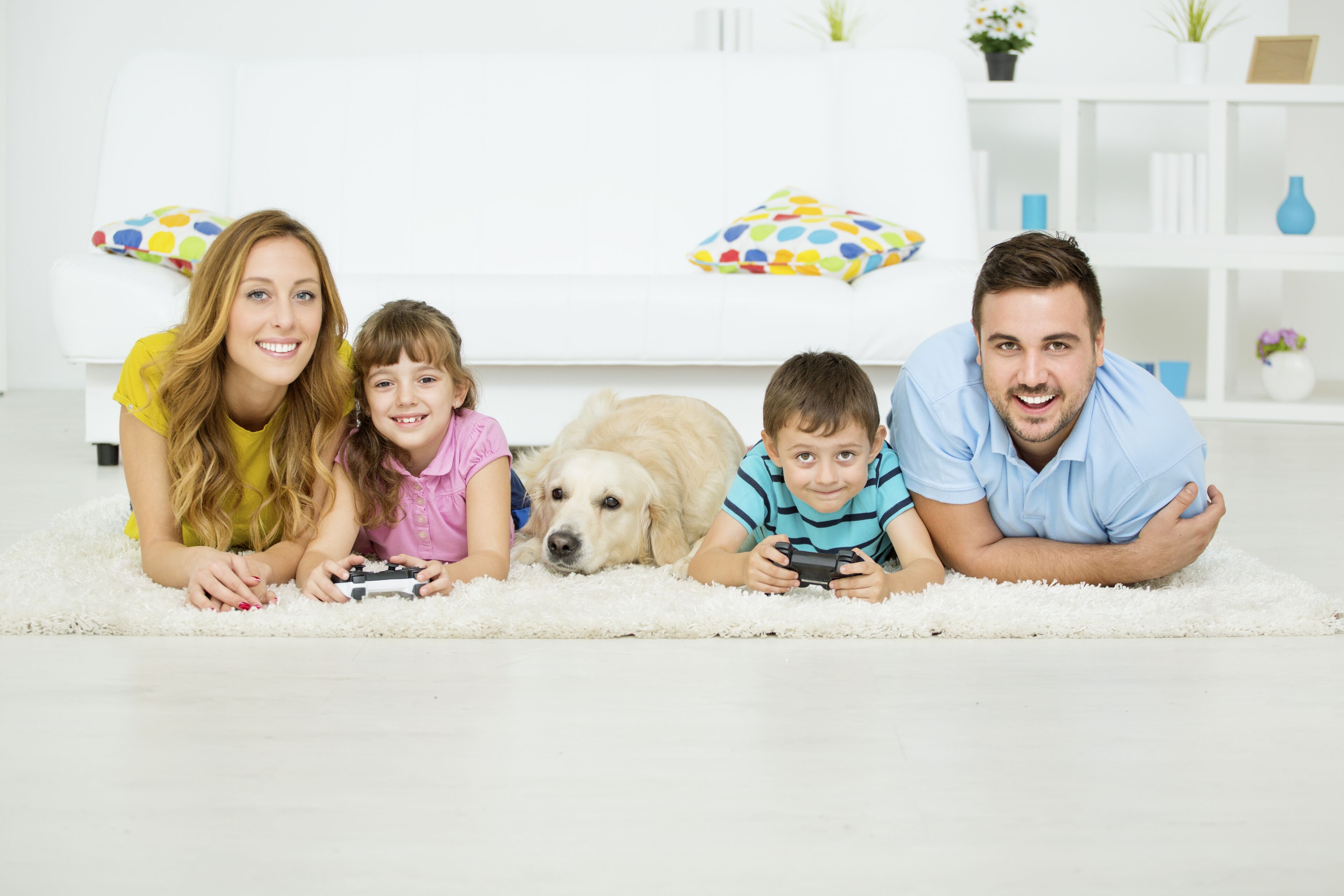 A family of four lying on a white carpet in a bright room with a white sofa and colorful pillows.