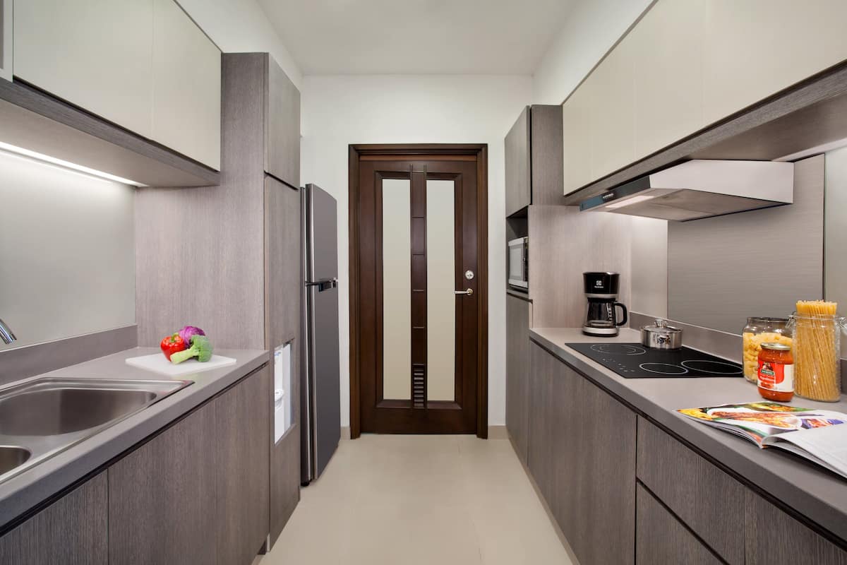 Une cuisine étroite équipée d’un évier en acier inoxydable, d’un four à micro-ondes, d’une machine à café et d’une cuisinière. Une porte donne accès à un garde-manger ou à un espace de rangement.