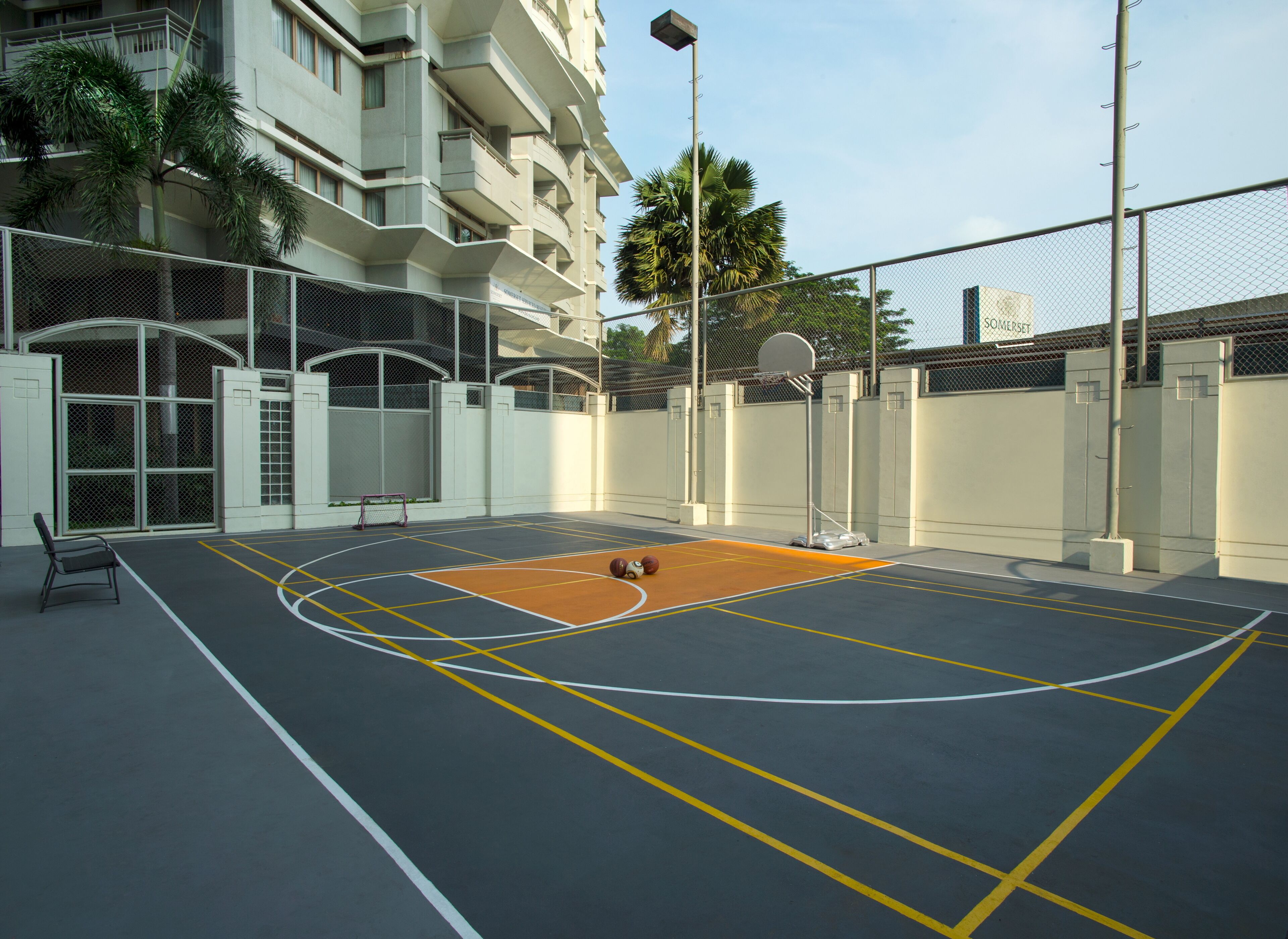 Een basketbalveld buiten, met een basketbalring, omheind door een hek en een gebouw op de achtergrond.