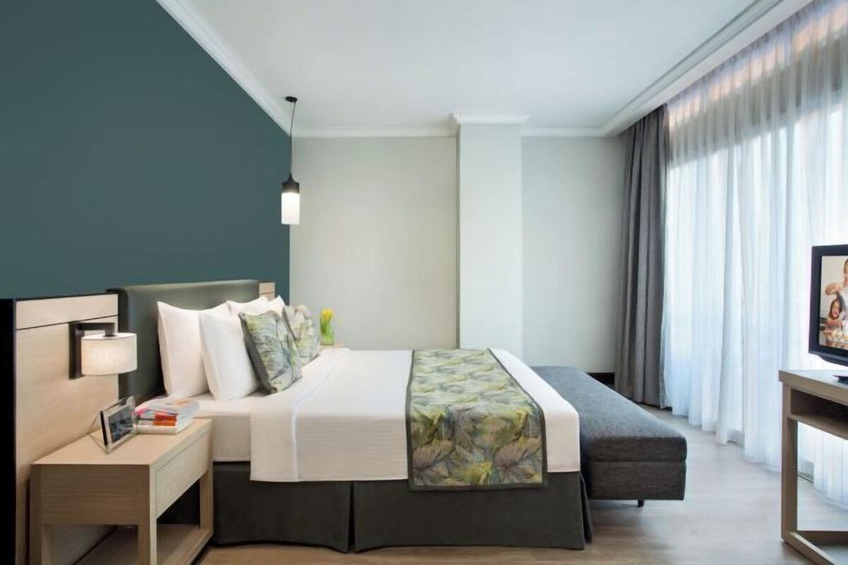 A modern hotel room with a bed, bedside table, lamp, and a view of the outdoors through large windows.