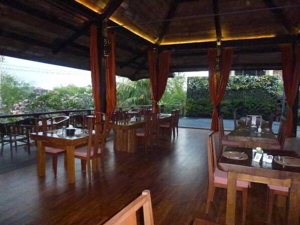 Un comedor interior con mesas y sillas de madera, un techo alto y vistas a zonas verdes al exterior.