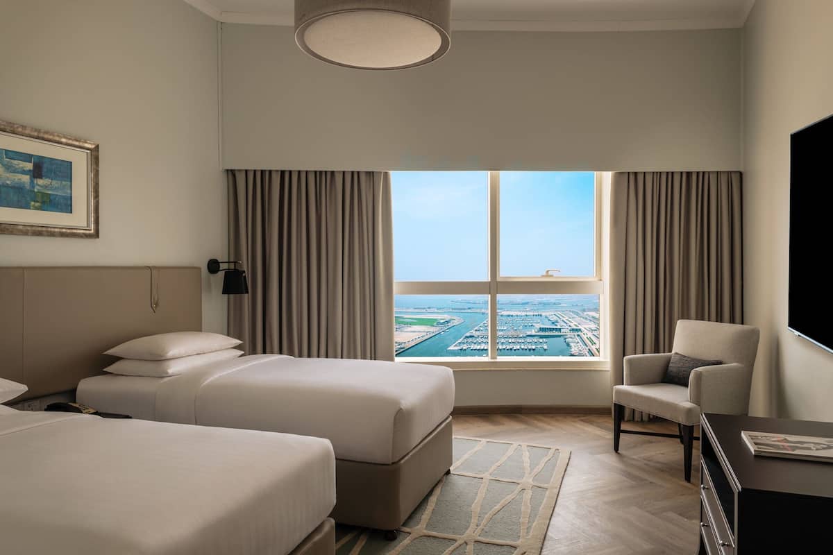 Habitación de hotel con dos camas, una silla, un escritorio y un ventanal con vistas a un puerto deportivo.