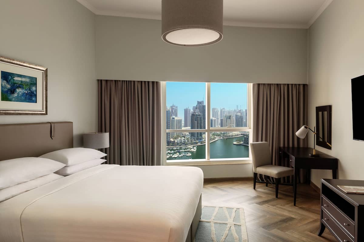 Habitación de hotel con una cama grande, un escritorio, una silla, una lámpara y vistas a un paisaje urbano a través de un ventanal.