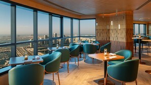 A modern restaurant with large windows offering city views, wooden accents, and teal chairs.