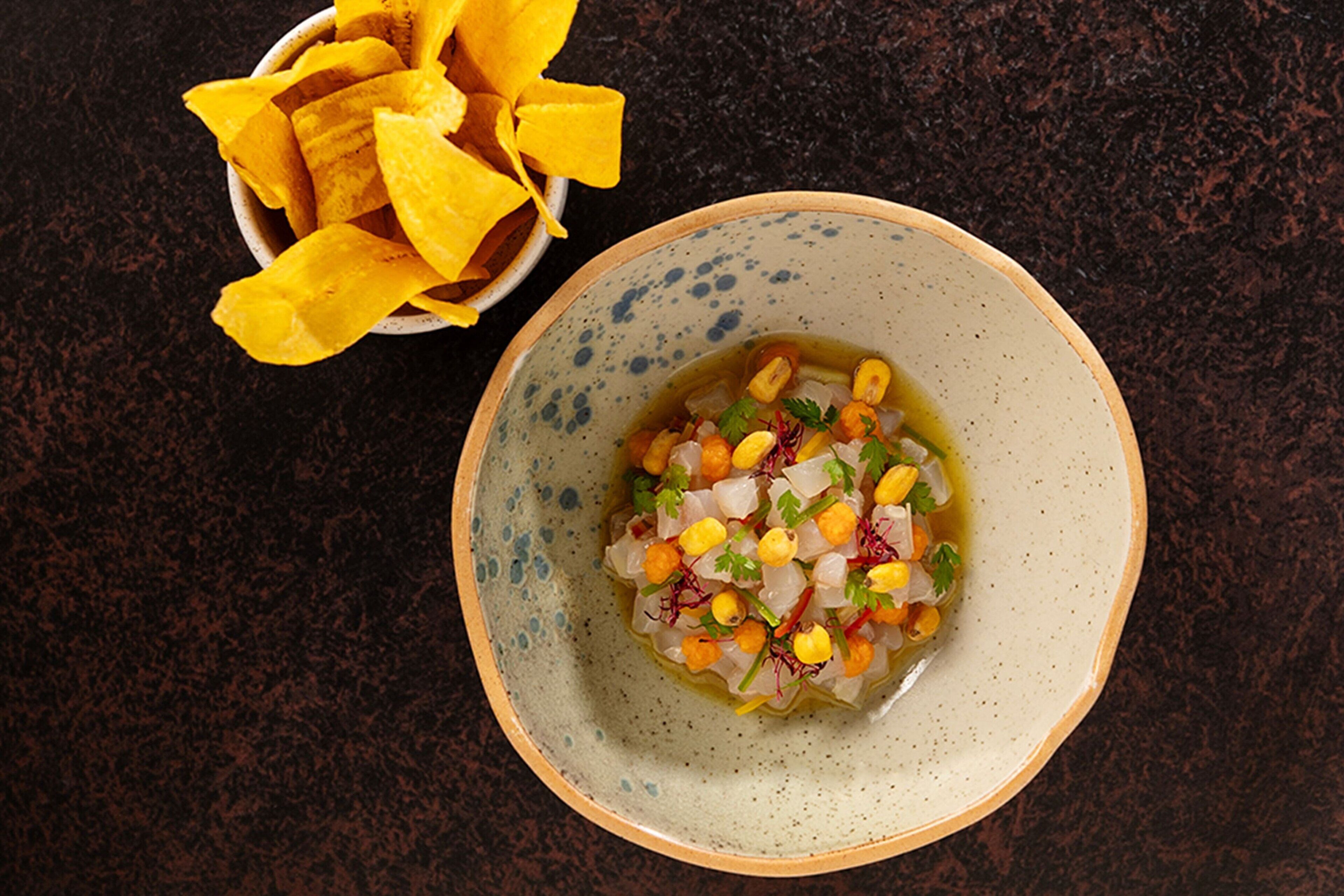 A bowl of ceviche with corn and onions, accompanied by a bowl of yellow corn chips on a dark surface.