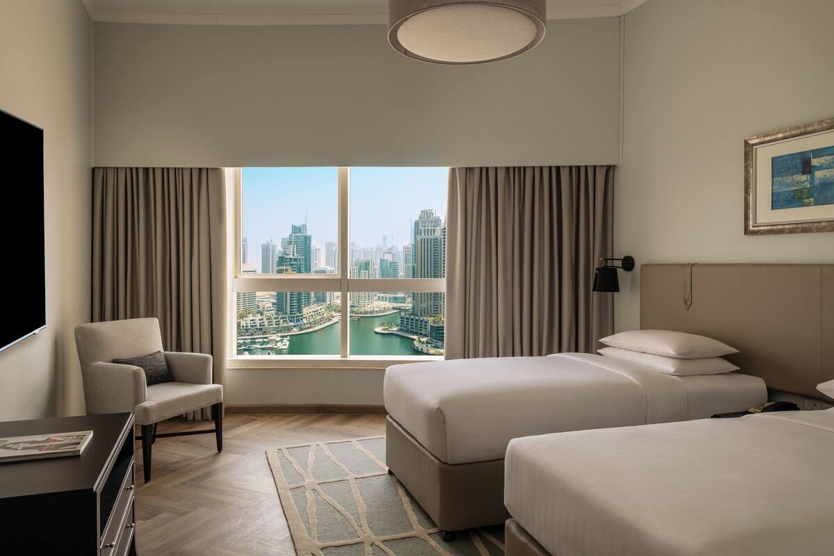Habitación de hotel con dos camas, una silla, un escritorio y un ventanal con vistas a la ciudad.