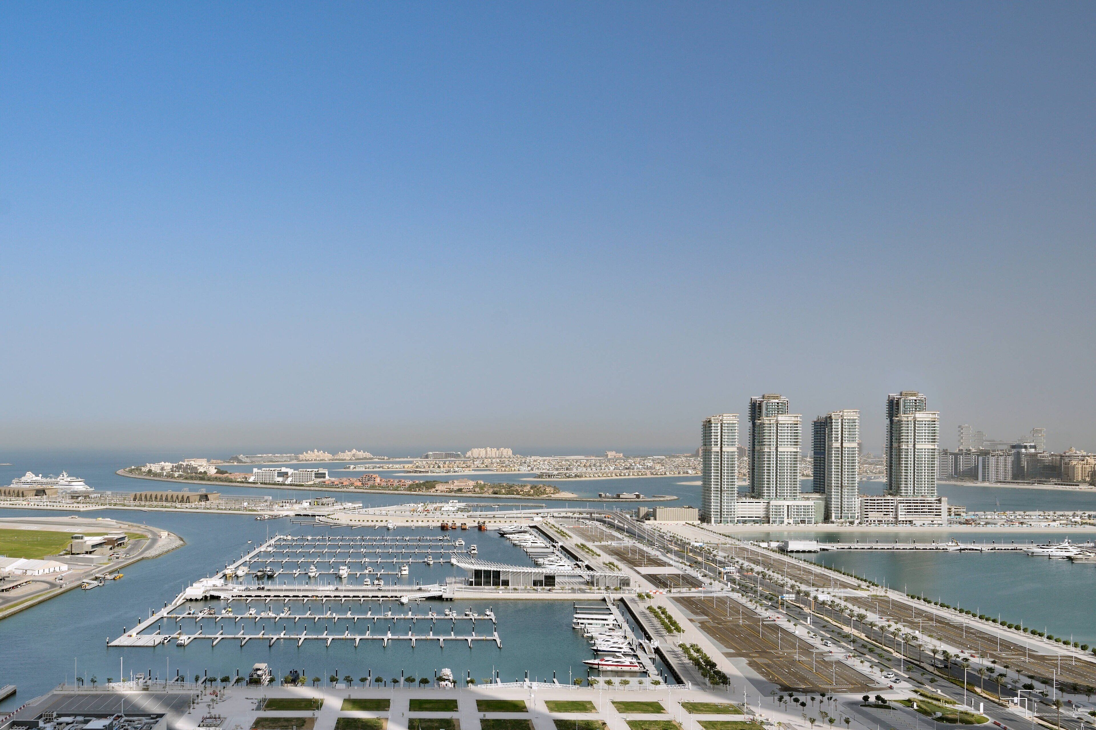 A marina with boats docked, a waterfront promenade, and high-rise buildings in the background.