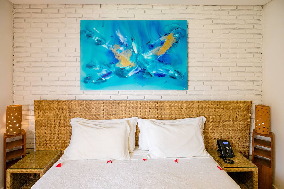 A hotel room with a white brick wall, a large abstract painting, a bed with white linens, two bedside tables with lamps, and a telephone on one of the tables.