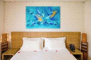 A hotel room with a white brick wall, a large abstract painting, a bed with white bedding, and a telephone on a woven table beside the bed. - Grand Oca Maragogi (Maragogi)