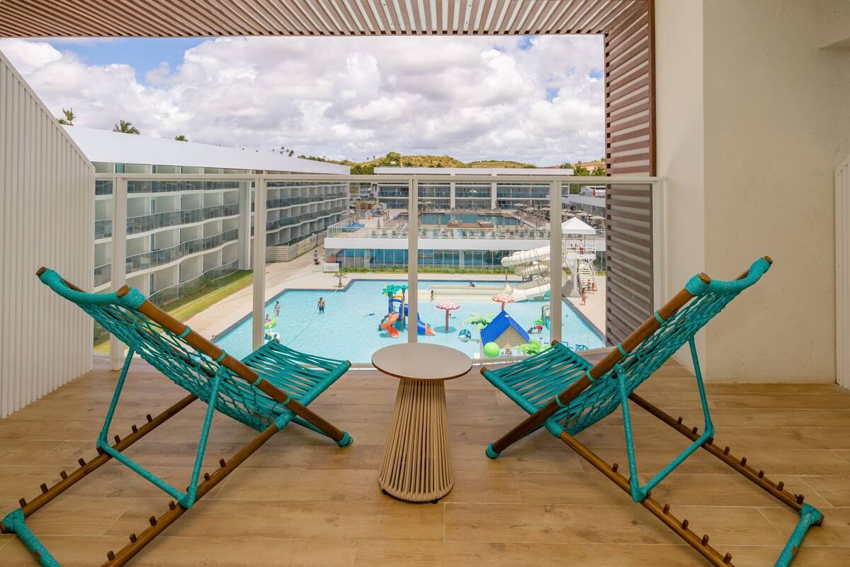 A balcony with two turquoise hammocks, a small round table, and a view of a pool area with a water slide.