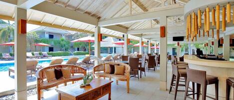 A covered outdoor lounge area with wooden furniture, a pool, and umbrellas.