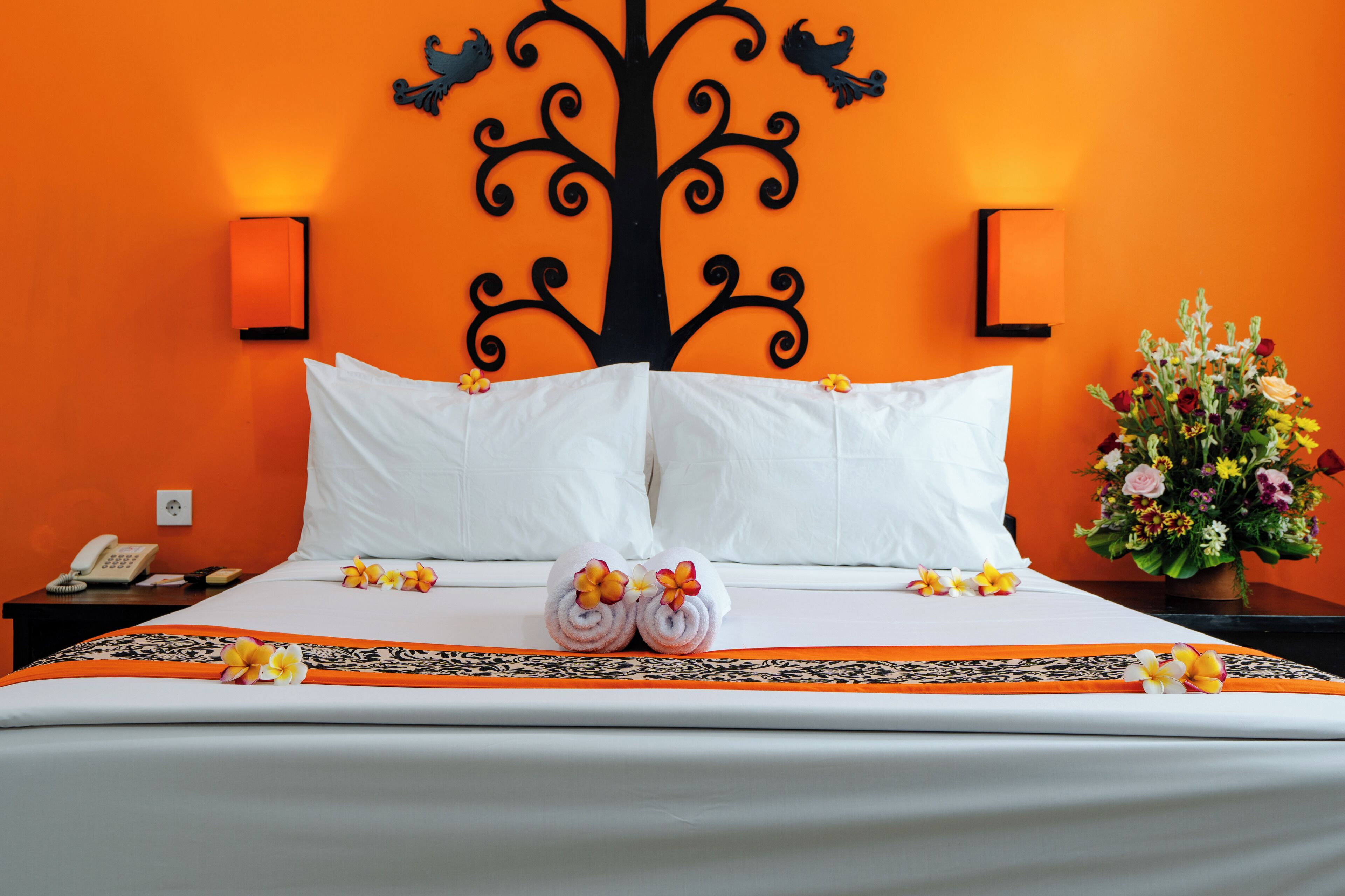 A hotel room with a large bed, white linens, a decorative tree wall art, two wall-mounted lamps, a nightstand with a phone, and a floral arrangement.