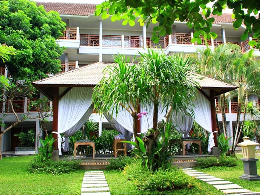 A hotel with a covered outdoor seating area, lush greenery, and a well-maintained garden.