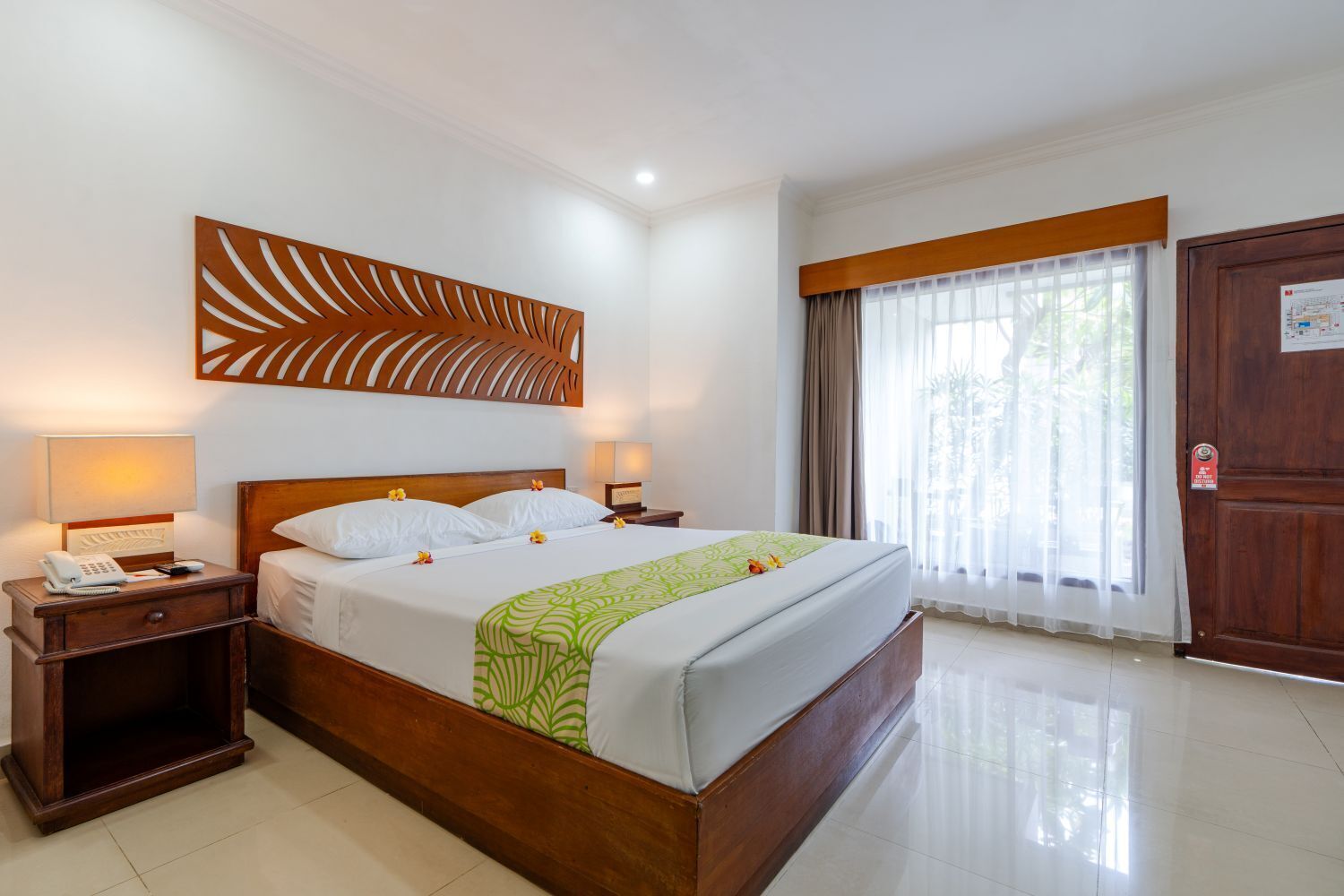Une chambre d’hôtel avec un grand lit, une tête de lit en bois, deux tables de chevet avec des lampes, un élément décoratif mural, une grande fenêtre avec des rideaux et une porte avec une pancarte.