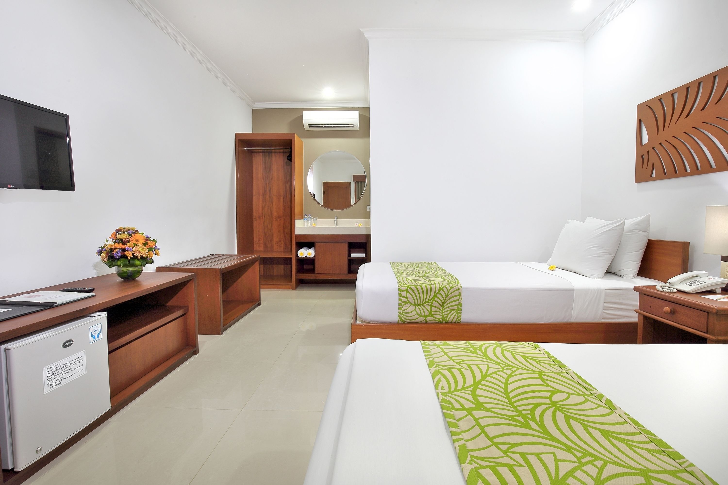 A hotel room with a bed, bedside table, a desk with a computer, a small fridge, a mirror, and a TV on the wall.