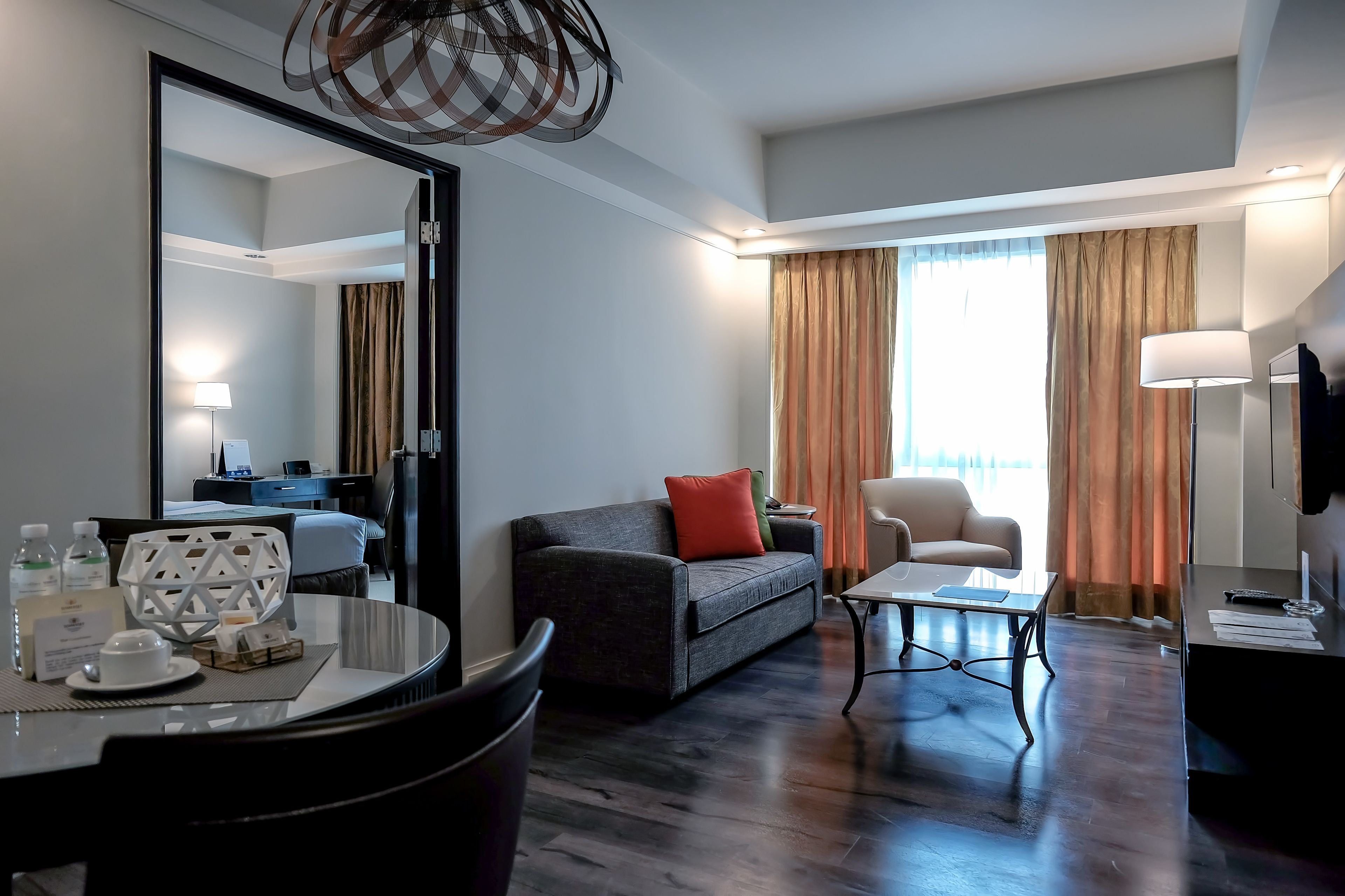 Ein modernes Hotelzimmer mit dunklem Holzboden, einem grauen Sofa mit rotem Kissen, einem weißen Sessel, einem kleinen runden Tisch und einem großen Spiegel.