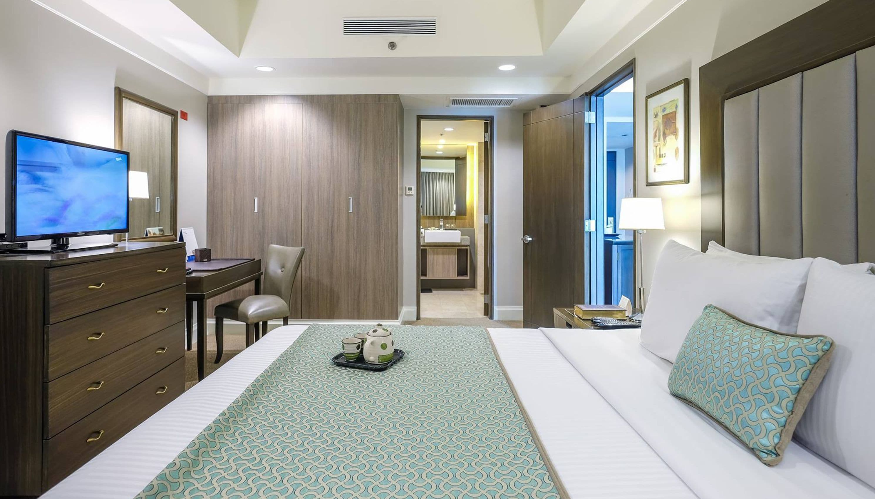 Habitación de hotel con una cama, un escritorio con computadora, una silla, un televisor y un baño visible a través de una puerta abierta.