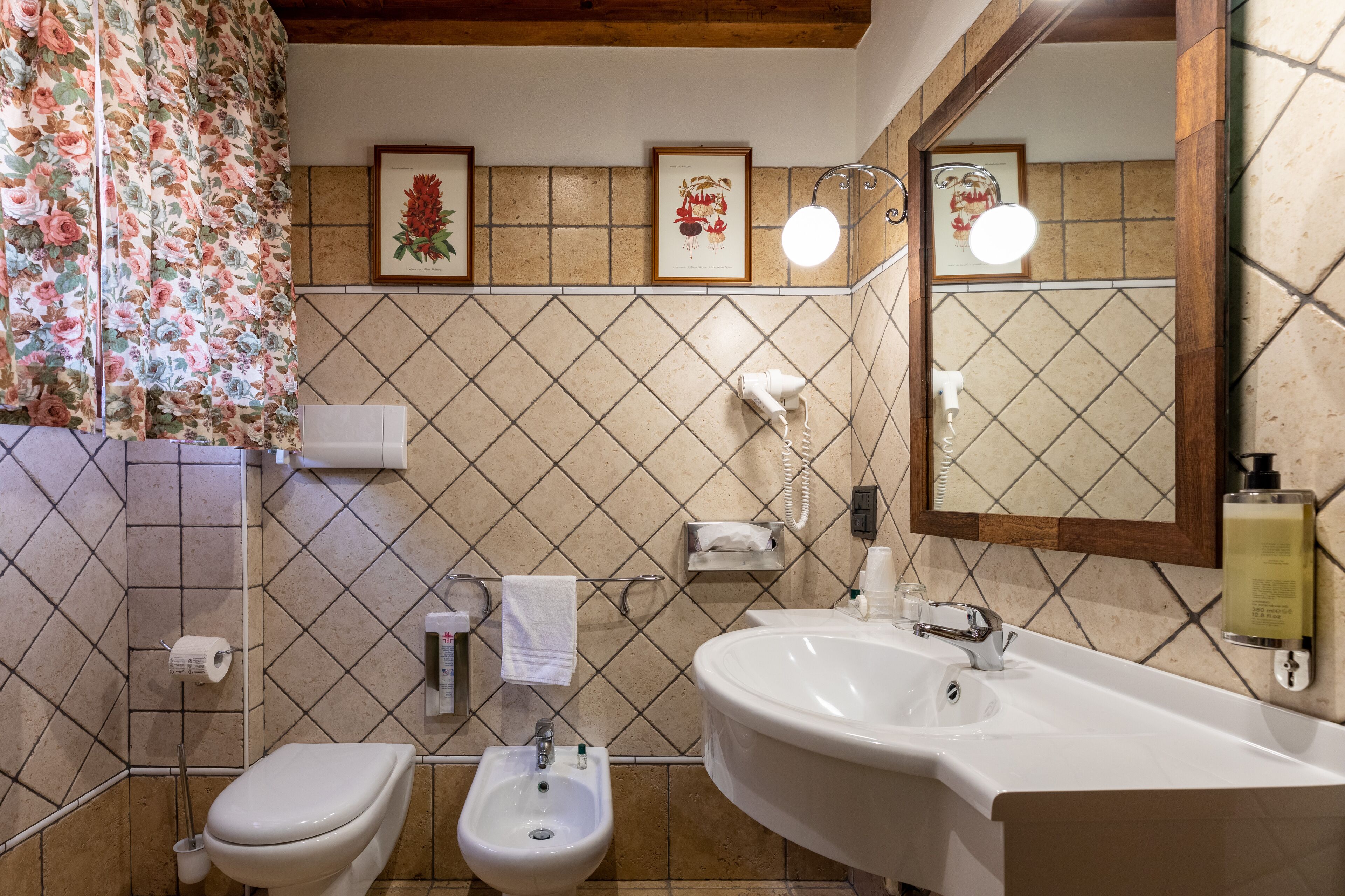 Un bagno con WC, bidet e lavandino, pareti piastrellate e quadri appesi.