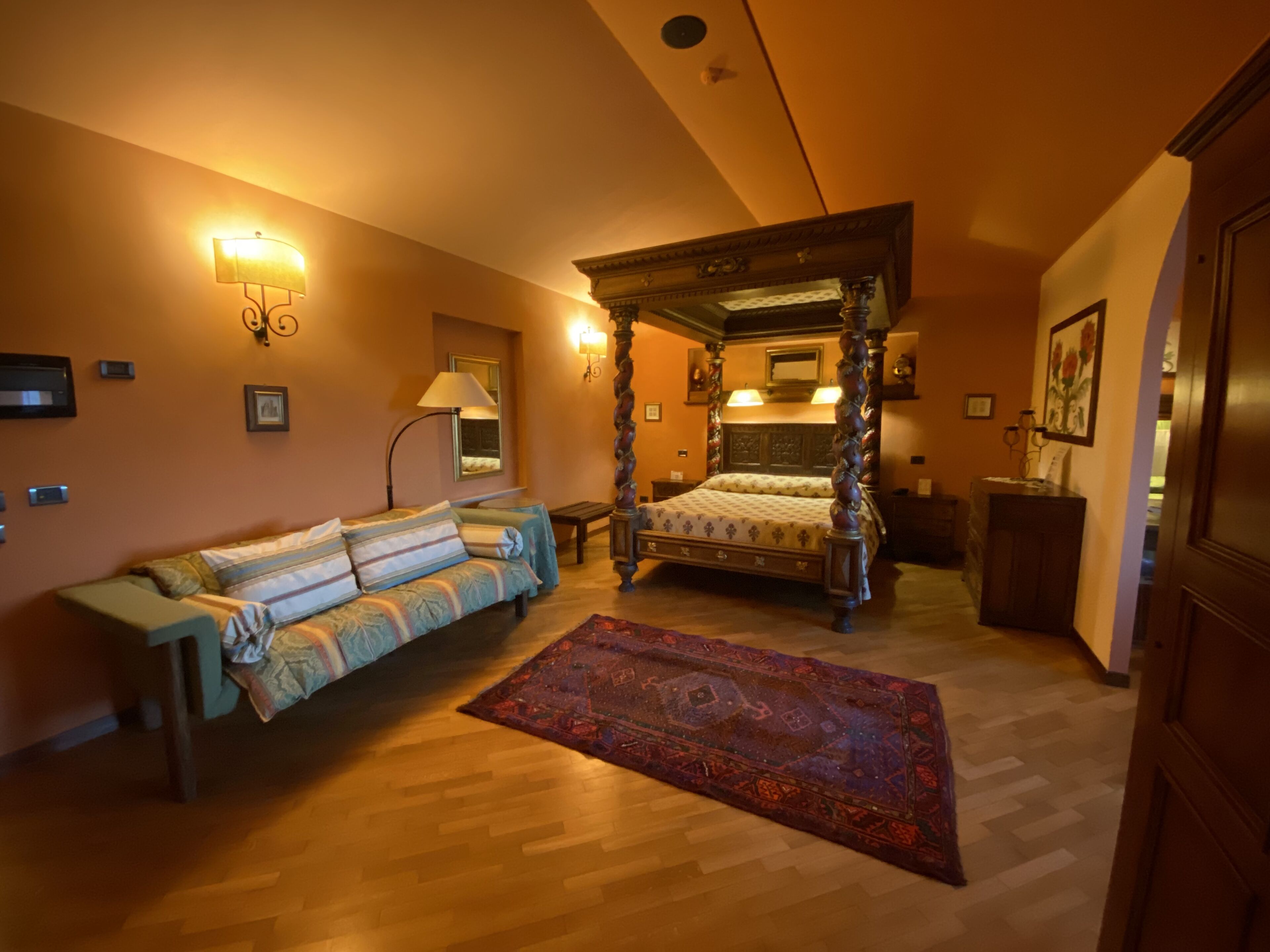 A bedroom with a four-poster bed, a sofa, a wooden floor, and wall-mounted lamps.