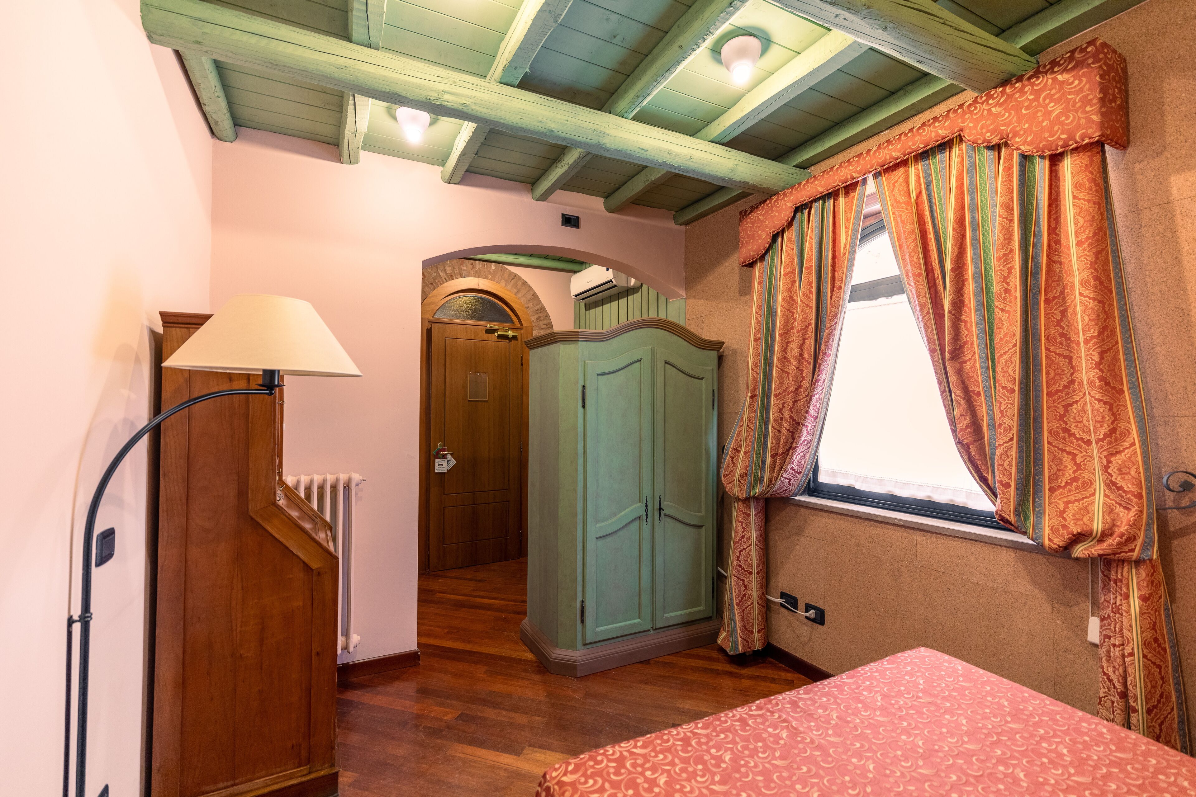 A room with a wooden floor, a bed with a patterned bedspread, a wardrobe, a lamp, and a window with curtains.