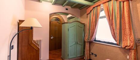 A room with a wooden floor, a bed with a patterned bedspread, a wardrobe, a lamp, and a window with curtains.
