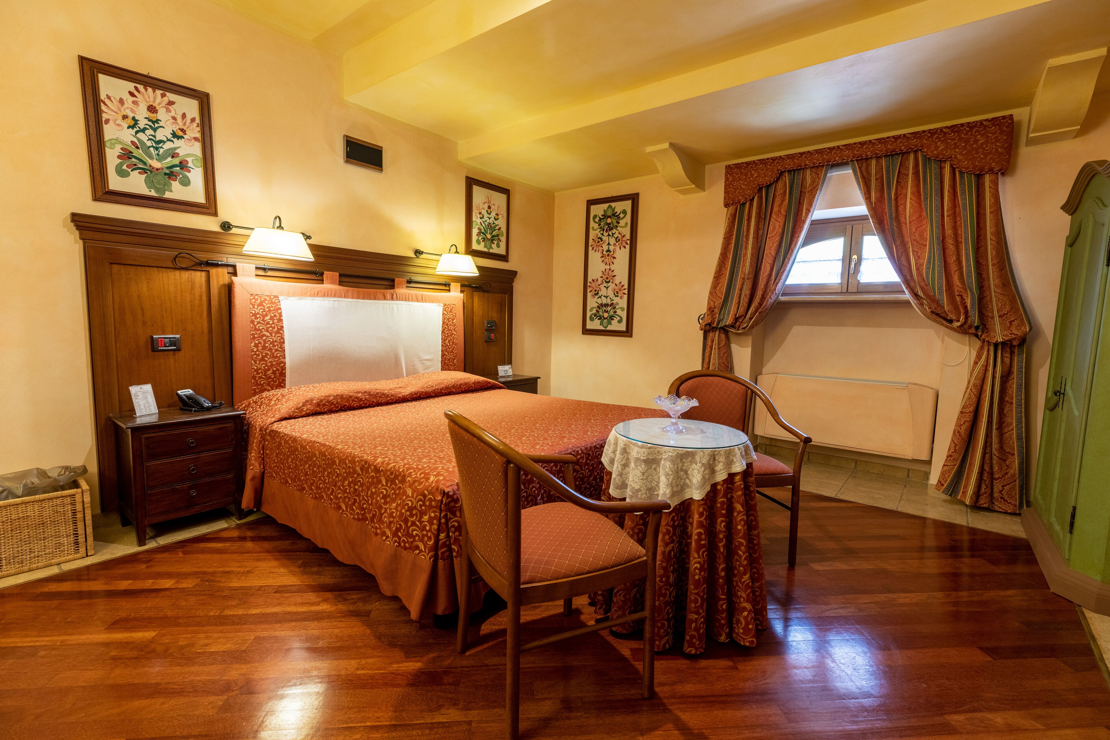 Habitación de hotel con piso de madera, una cama con colcha naranja, una mesita redonda pequeña con una silla, una mesita de noche con lámpara y una ventana con cortinas.