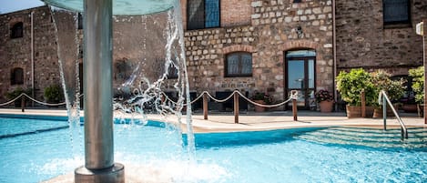 A swimming pool with a water fountain in the center, surrounded by a stone building with arched windows and a door.