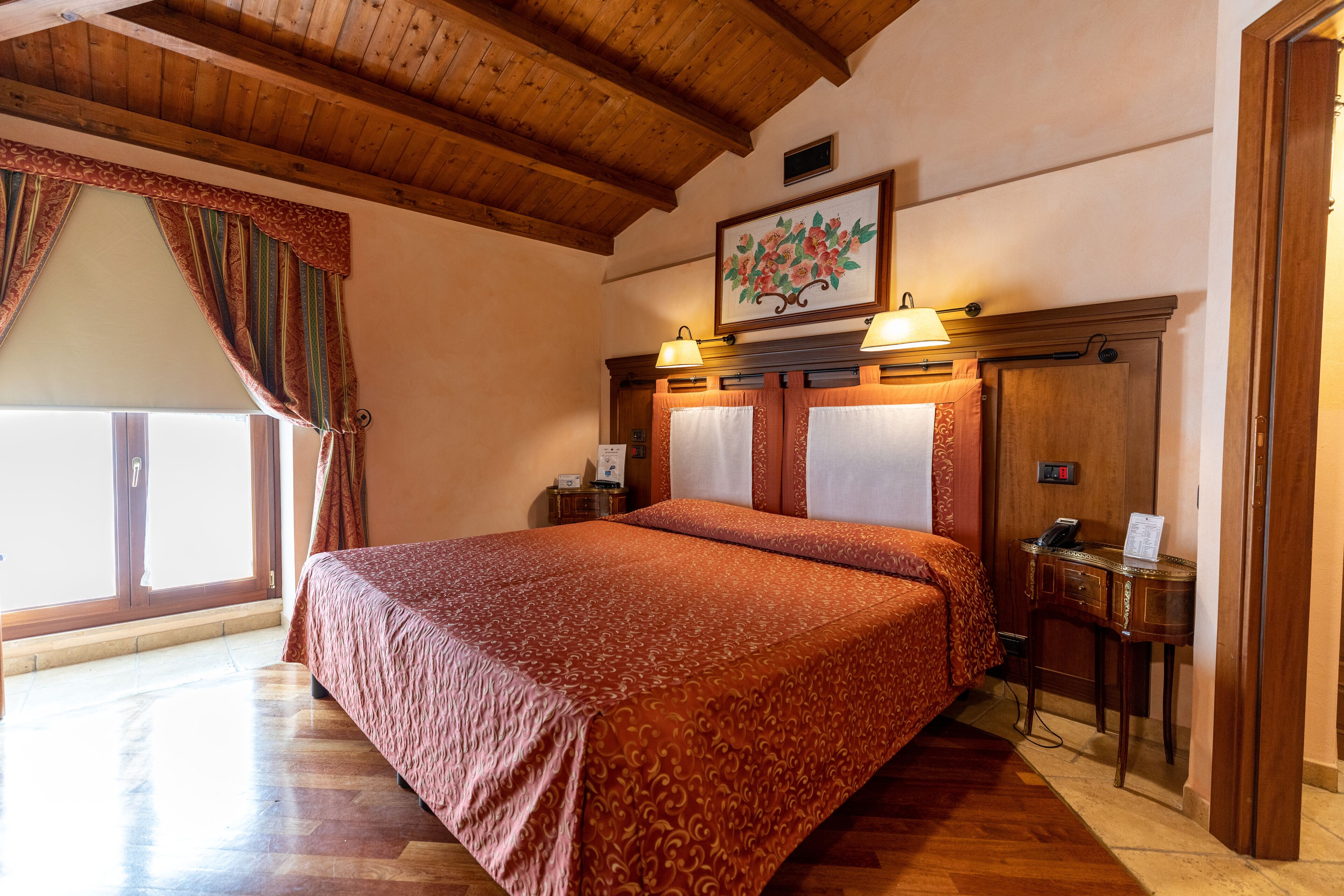 A hotel room with a large bed, wooden ceiling beams, a painting on the wall, and a desk with a lamp.