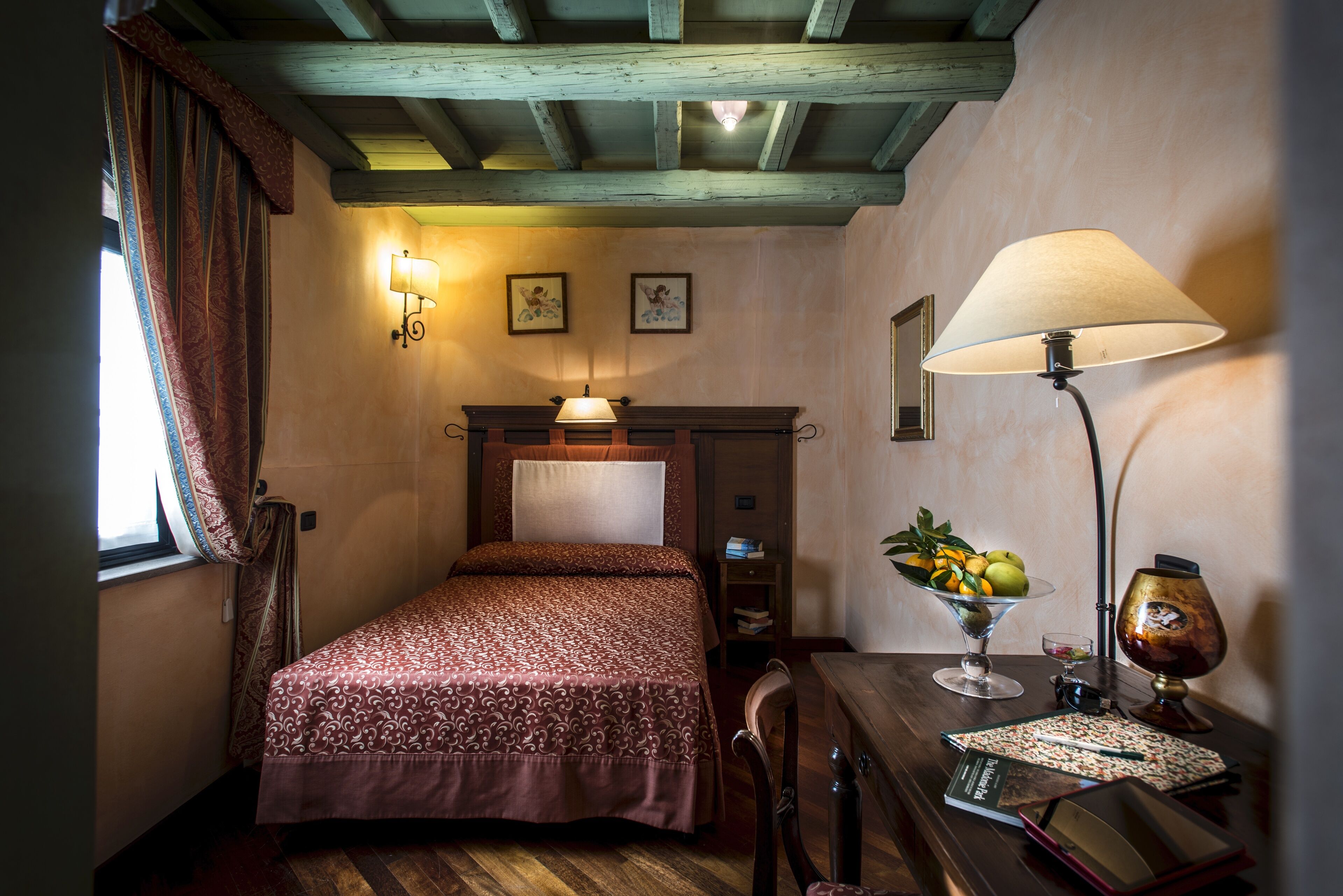 A bedroom with a bed, a desk with a lamp, a fruit bowl, and a wall-mounted shelf.