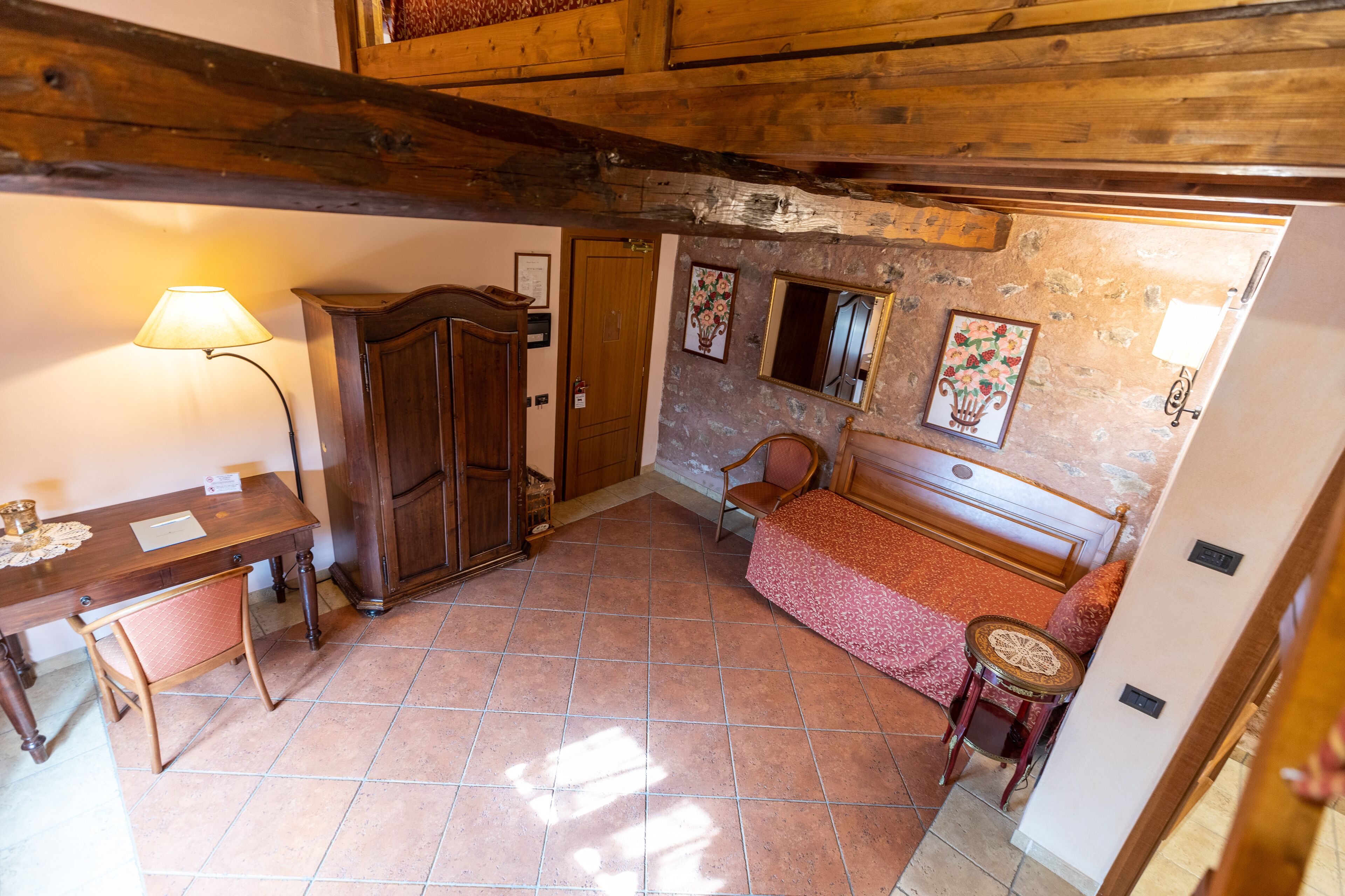 A room with a wooden ceiling, a bed with a red bedspread, a chair, a table with a lamp, and a wooden cabinet.