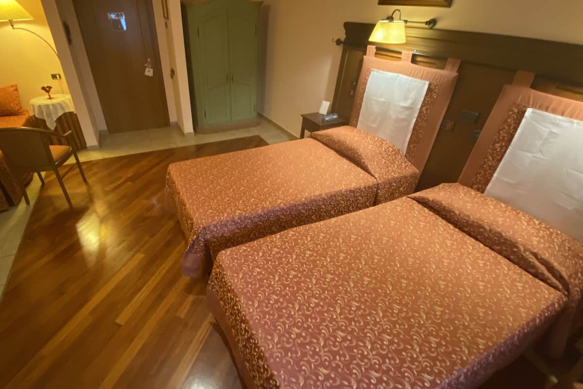 A hotel room with two beds, a small table, a chair, a nightstand, and a green door.