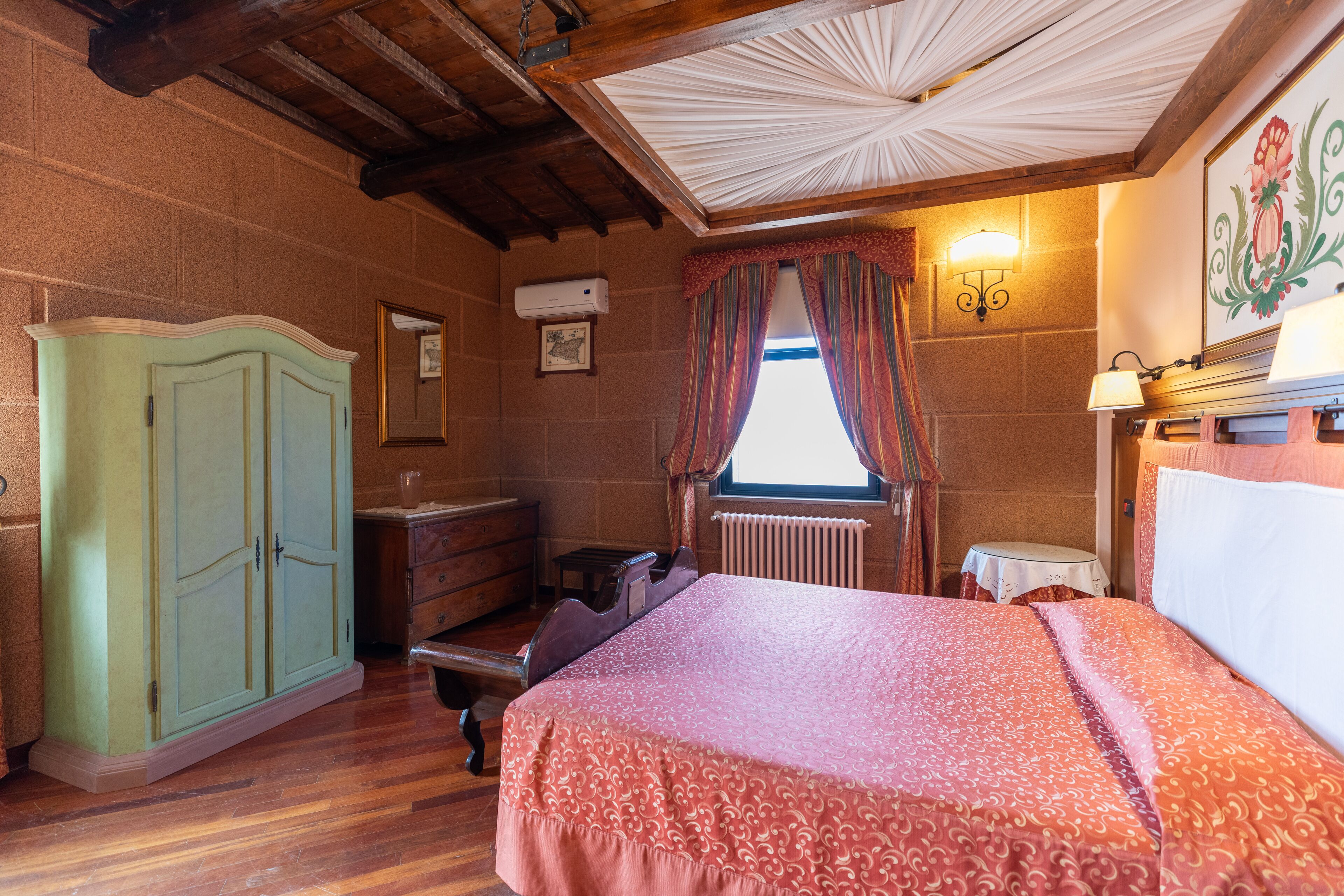 Una camera da letto con soffitto in legno, un letto con copriletto decorato, un armadio, una cassettiera, un termosifone e una finestra con tende.