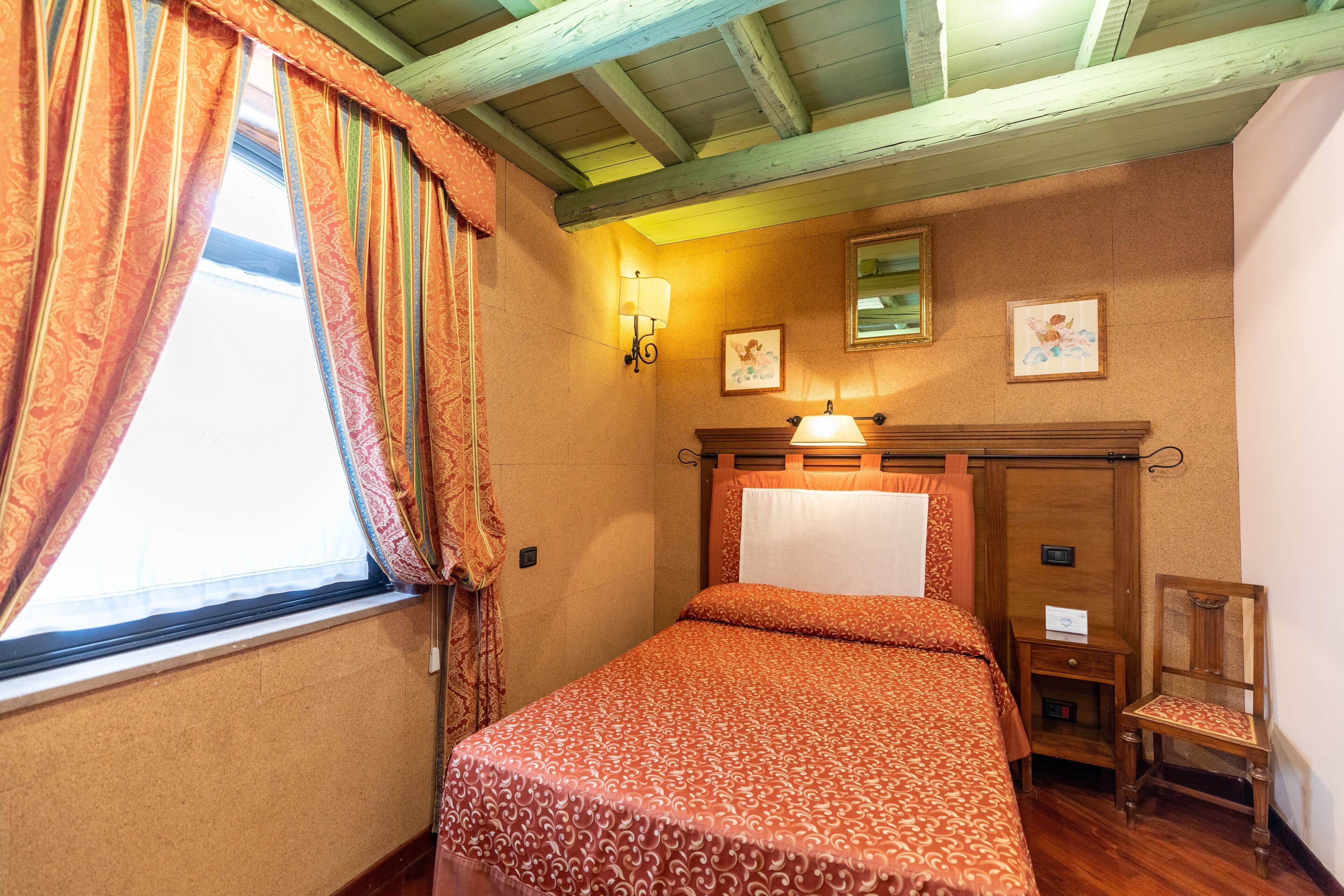 Un dormitorio con cabecera de madera, una cama individual, una mesita de noche, una silla y un ventanal con cortinas.