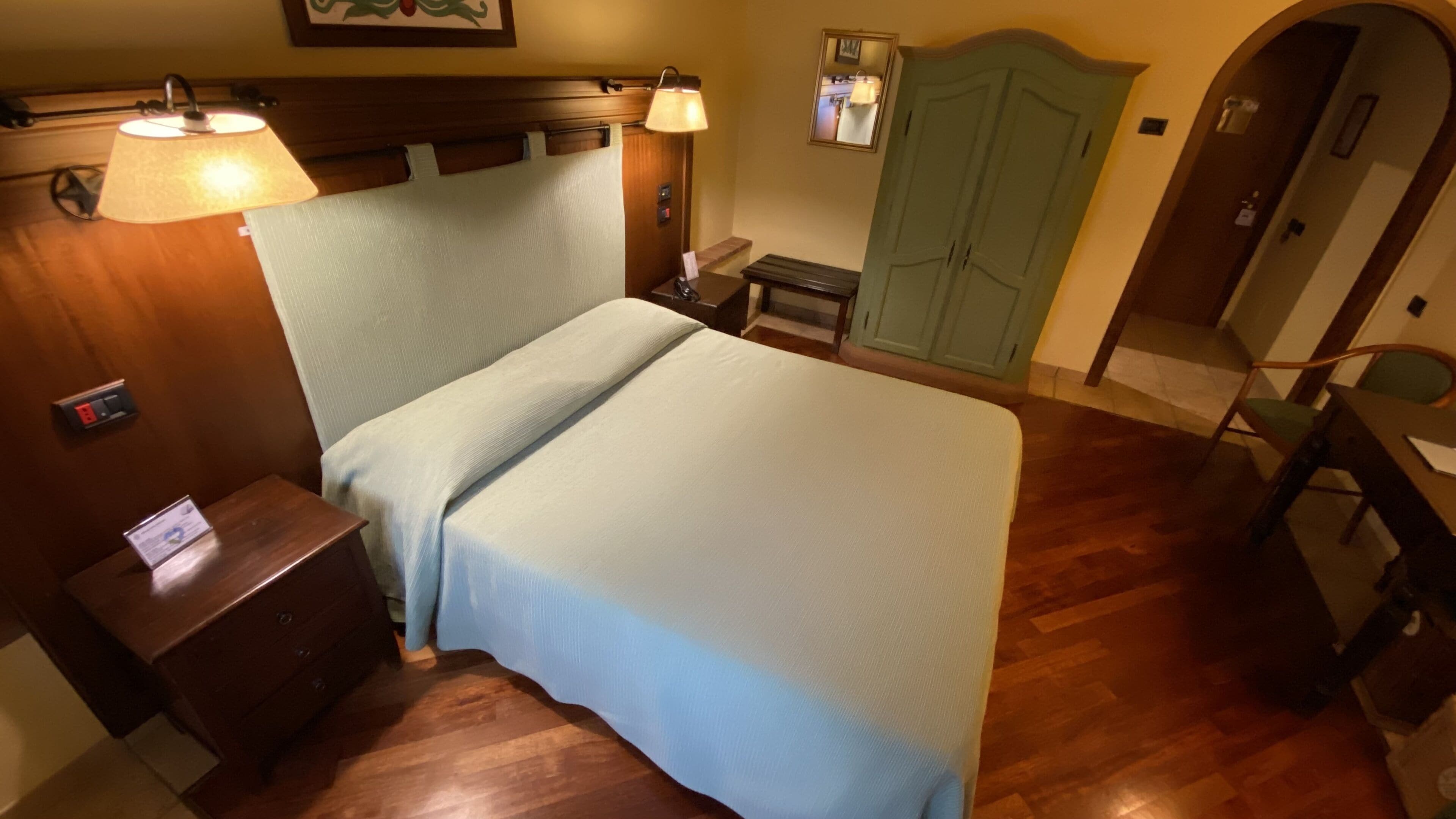 A hotel room with a bed, bedside table, lamp, wardrobe, and a desk with a chair.