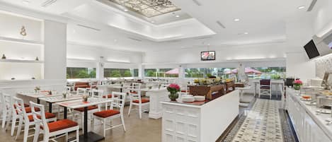 Un restaurante espacioso con mobiliario blanco, una isla central y un amplio ventanal con vistas a la vegetación.
