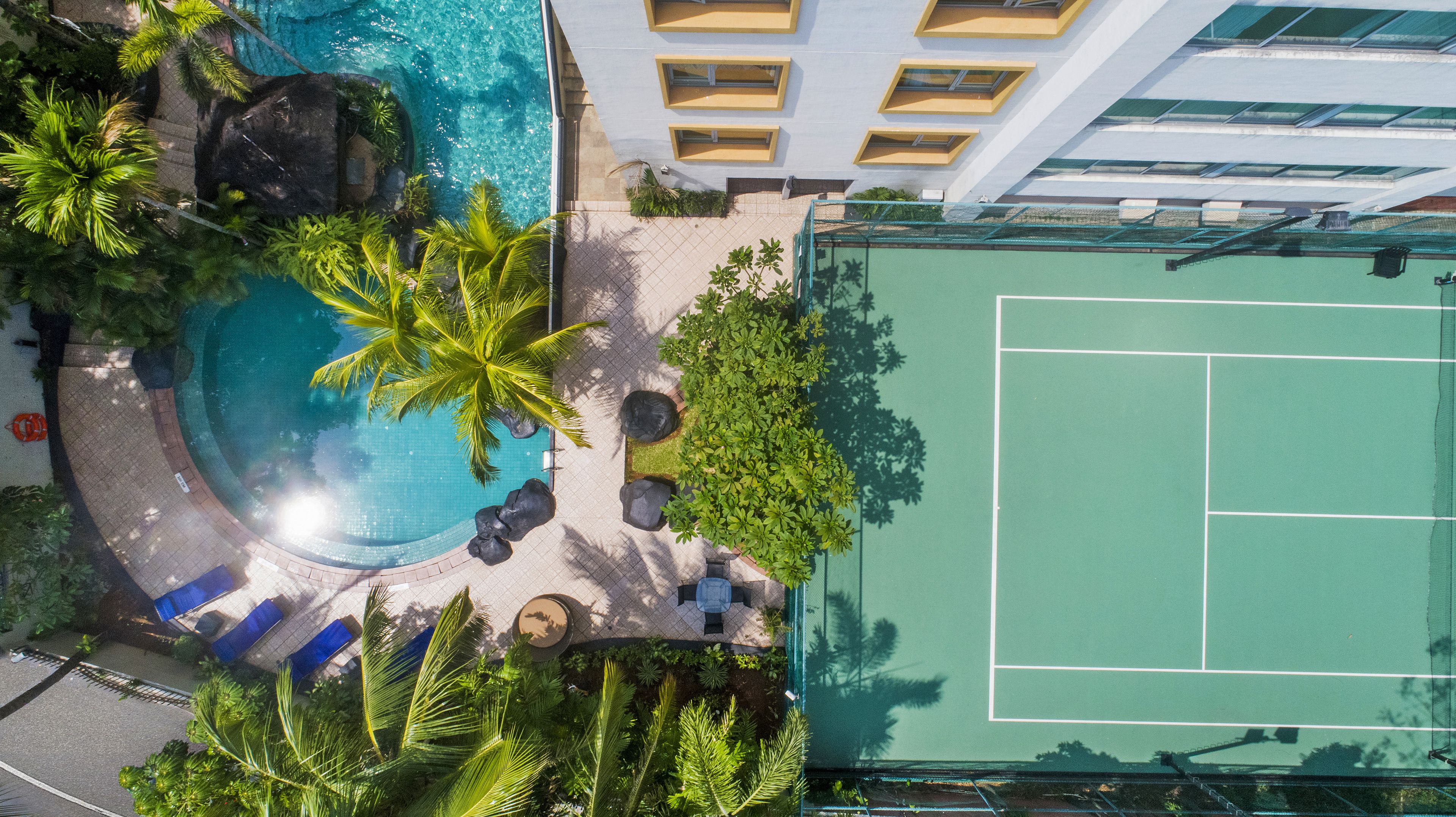 Vista aérea de un hotel con piscina, rodeado de palmeras y una cancha de tenis.