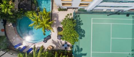 Vista aérea de un hotel con piscina, rodeado de palmeras y una cancha de tenis.