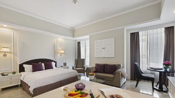 A hotel room with a large bed, a dining table with a book and a fruit platter, a sofa, a chair, and a small table with a vase. - Ascott Jakarta (Jakarta)