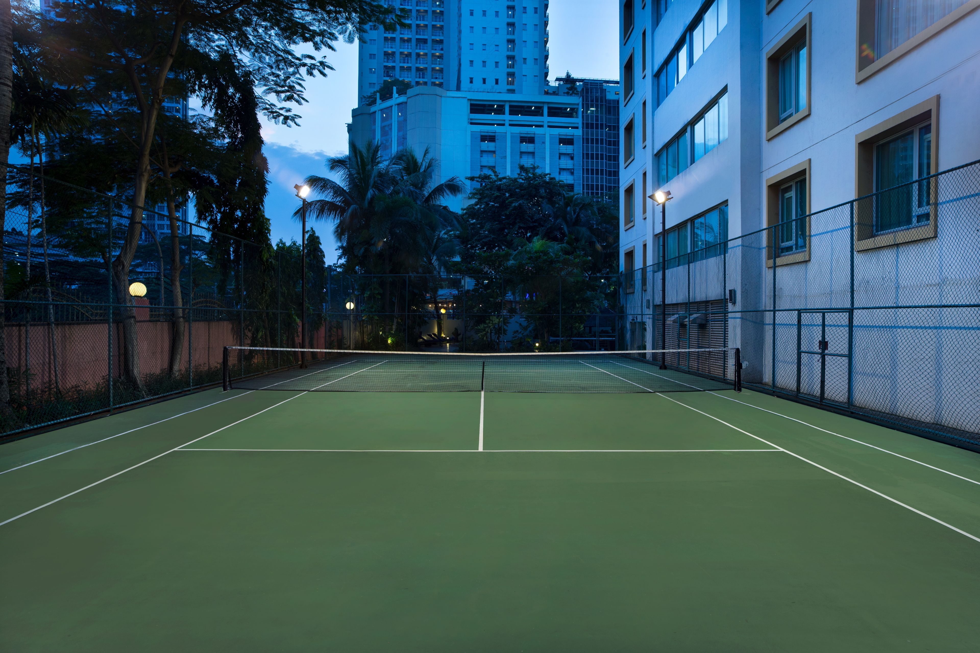 An indoor tennis court with a net, surrounded by a fence, and adjacent to a building with large windows and balconies.