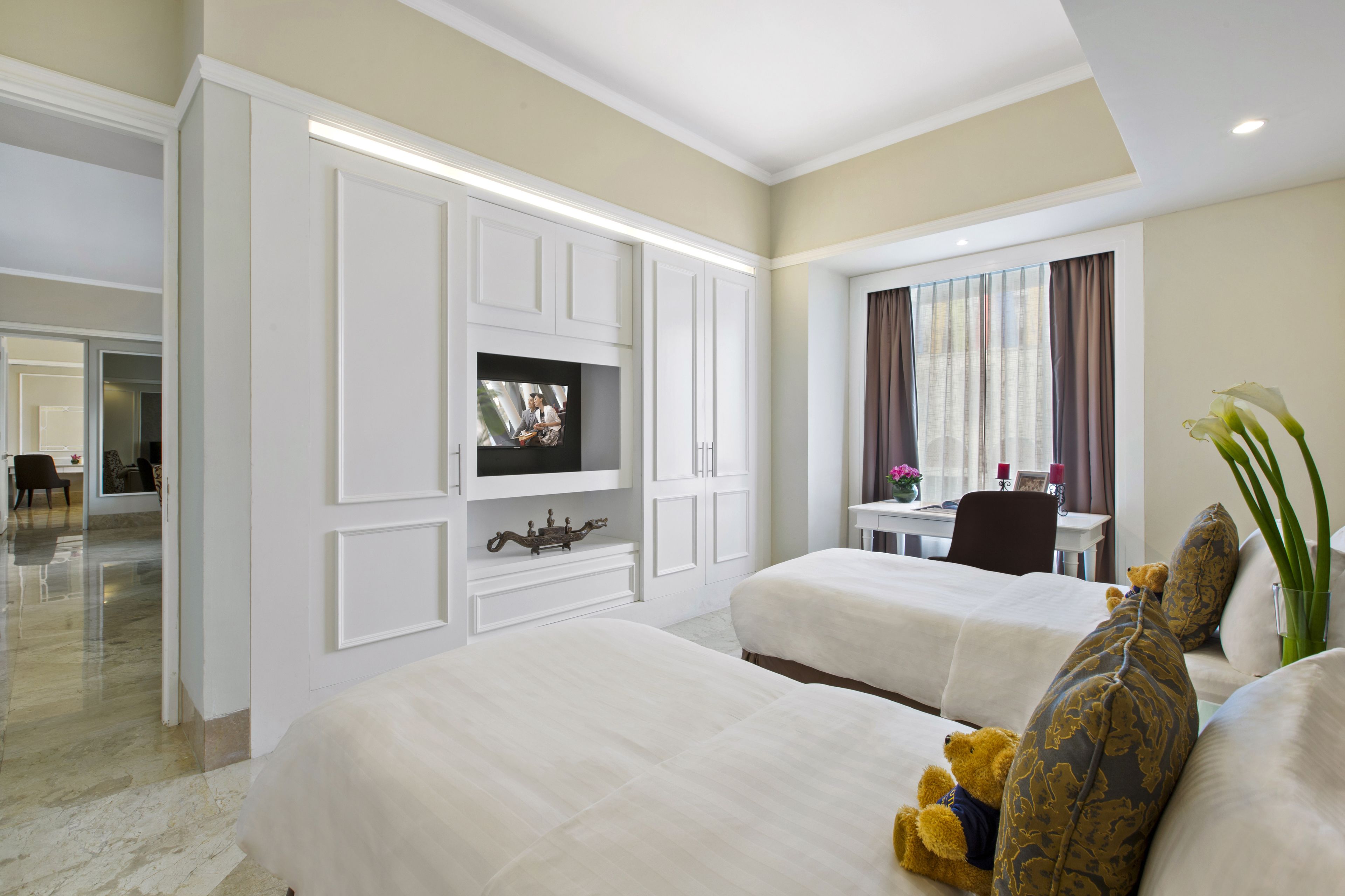 A hotel room with two beds, a TV, a desk, and a window with curtains.