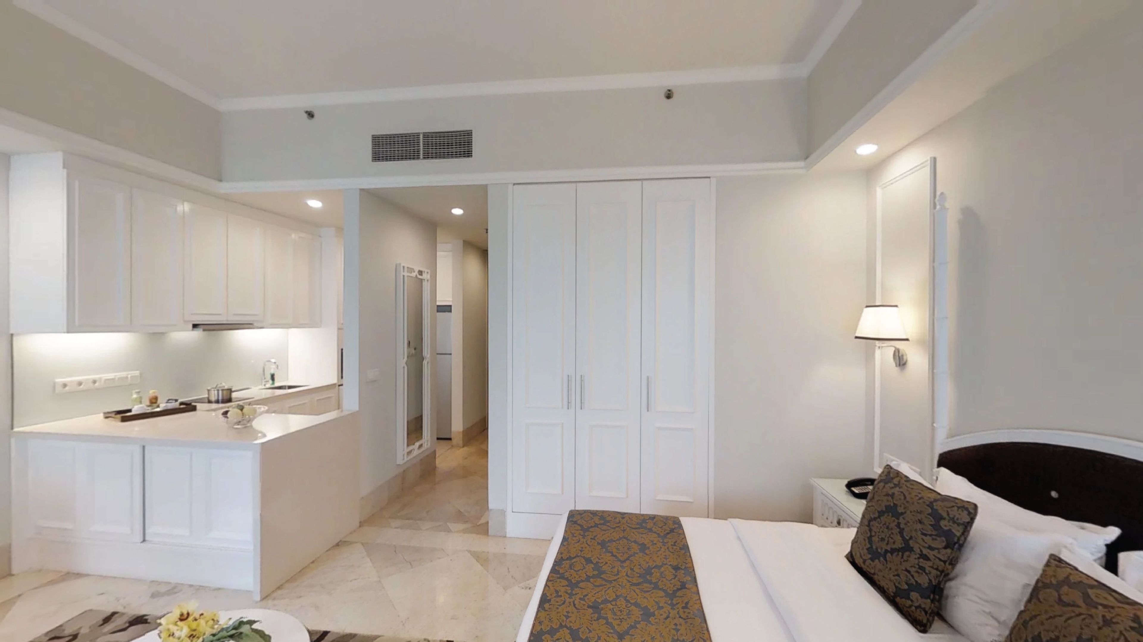 A hotel room with a bed, bedside table, lamp, and a view of the kitchen and hallway.