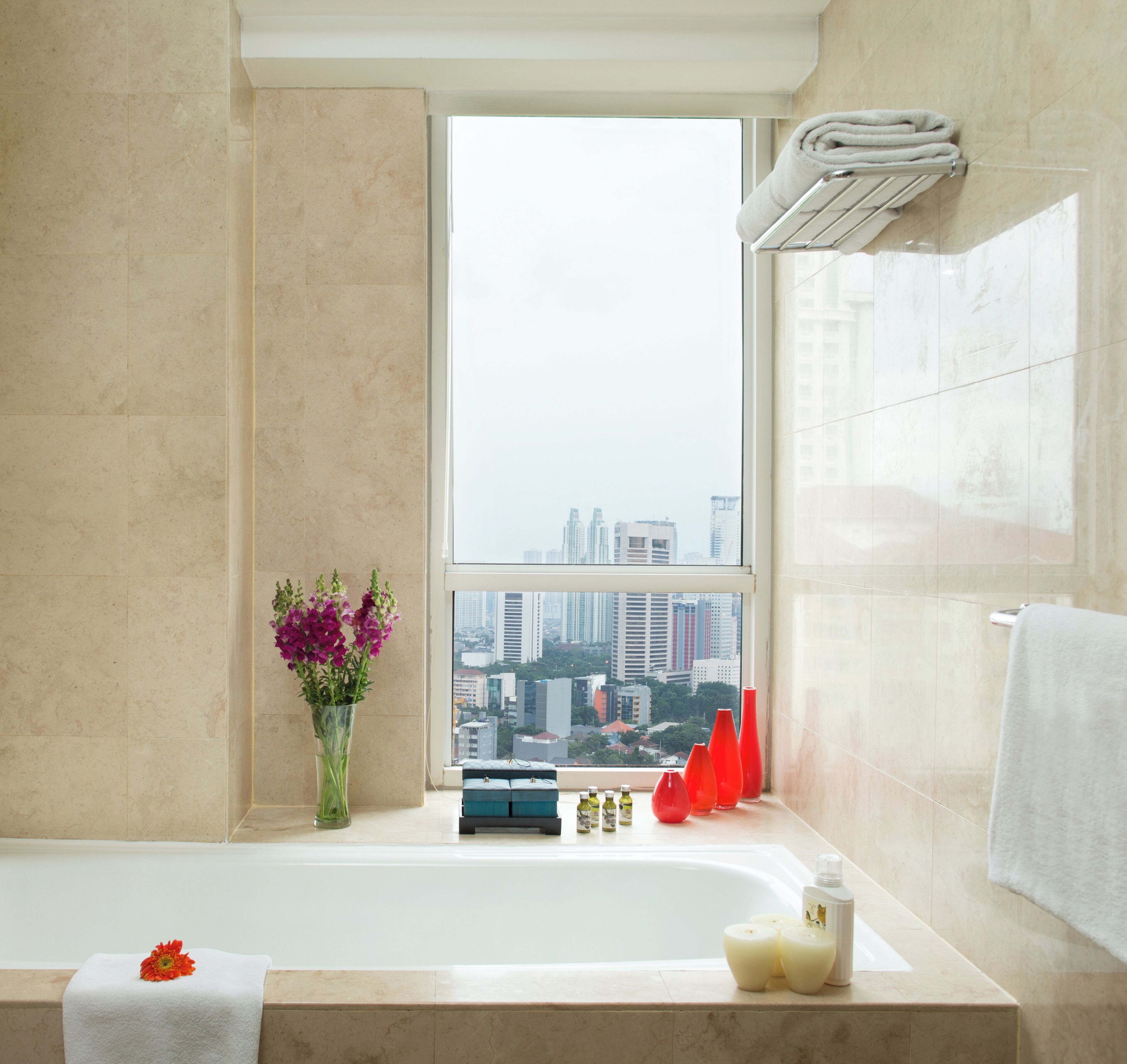 A modern bathroom with a bathtub, a window with a city view, a vase with flowers, and toiletries on the windowsill.