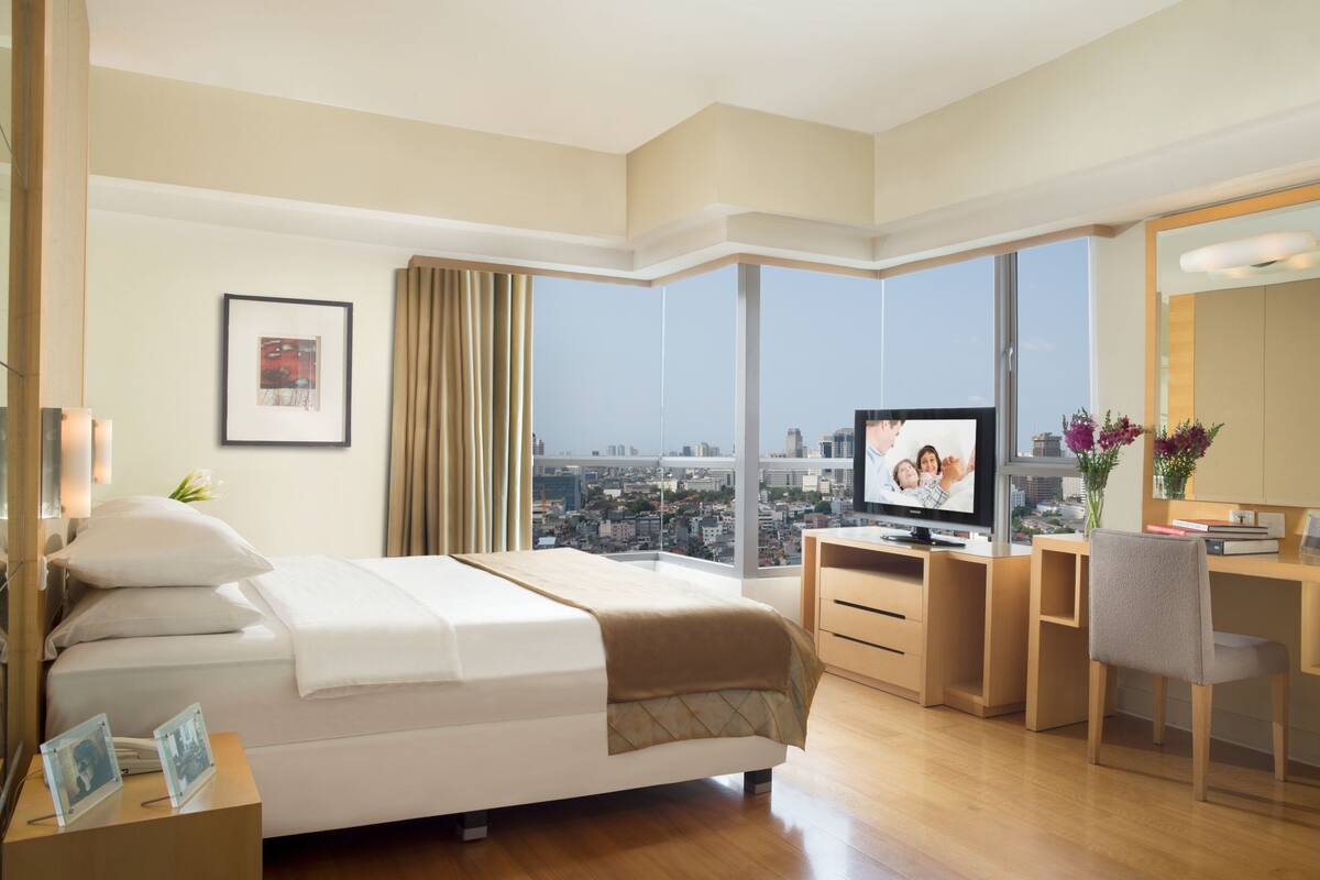 Een hotelkamer met een bed, een bureau, een televisie en een groot raam met uitzicht op de stad.