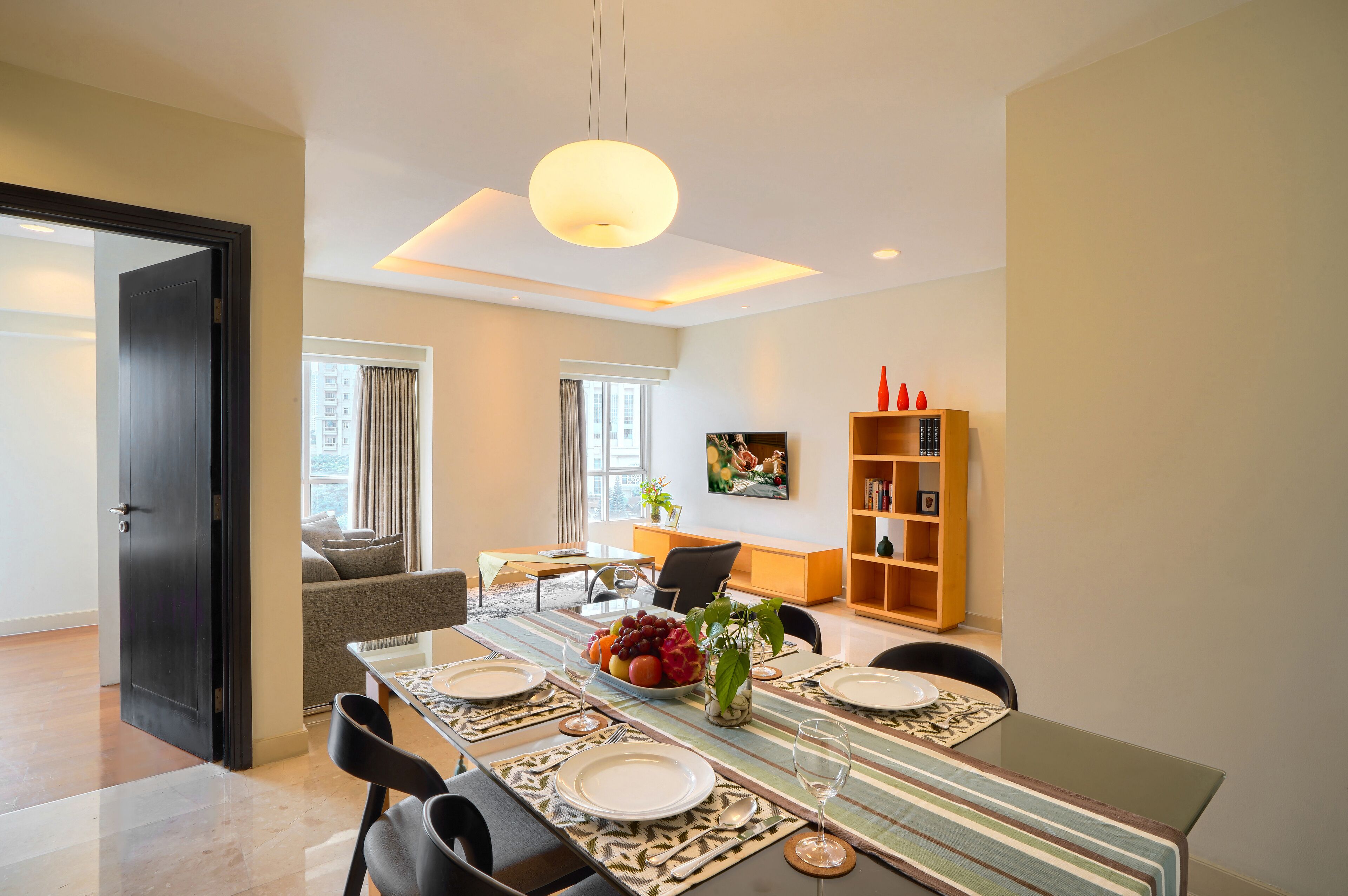 Uma sala de jantar moderna com uma mesa comprida posta para quatro pessoas, um sofá, uma TV e uma estante de livros.